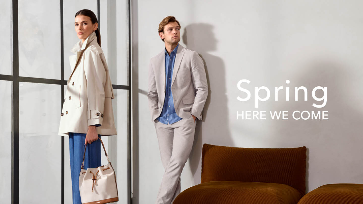 Zwei Models präsentieren Frühlingsmode: Eine Frau trägt einen beigen Mantel und hält eine Tasche, während ein Mann in einem grauen Anzug mit blauem Hemd posiert; daneben steht der Text 'Spring Here We Come'.
