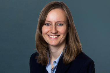 Porträt einer lächelnden Person mit schulterlangem braunem Haar in einem dunklen Blazer und hellen Hemd vor einem neutralen Hintergrund.