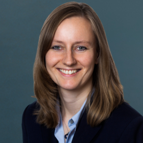 Porträt einer lächelnden Frau mit schulterlangem braunem Haar und einem dunkelblauen Blazer vor einem grauen Hintergrund.