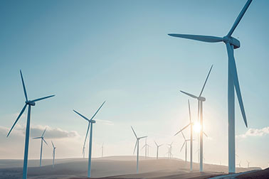 Wind turbines on a hilly landscape against a clear sky with the sun shining through.