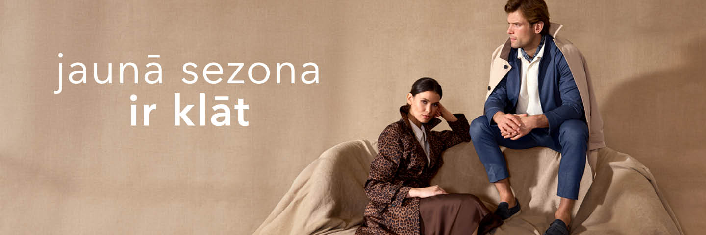 Двое моделей в стильной одежде сидят на диване на фоне бежевого цвета; текст на латышском языке — "jaunā sezona ir klāt".